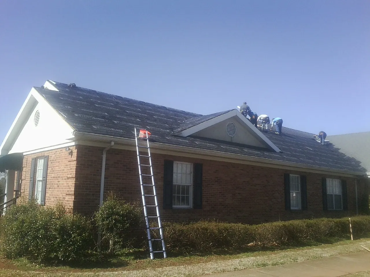 Crew tearing shingles off a brick ranch roof with a ladder set — Emergency Roof Repair work by our crew in Lake Geneva