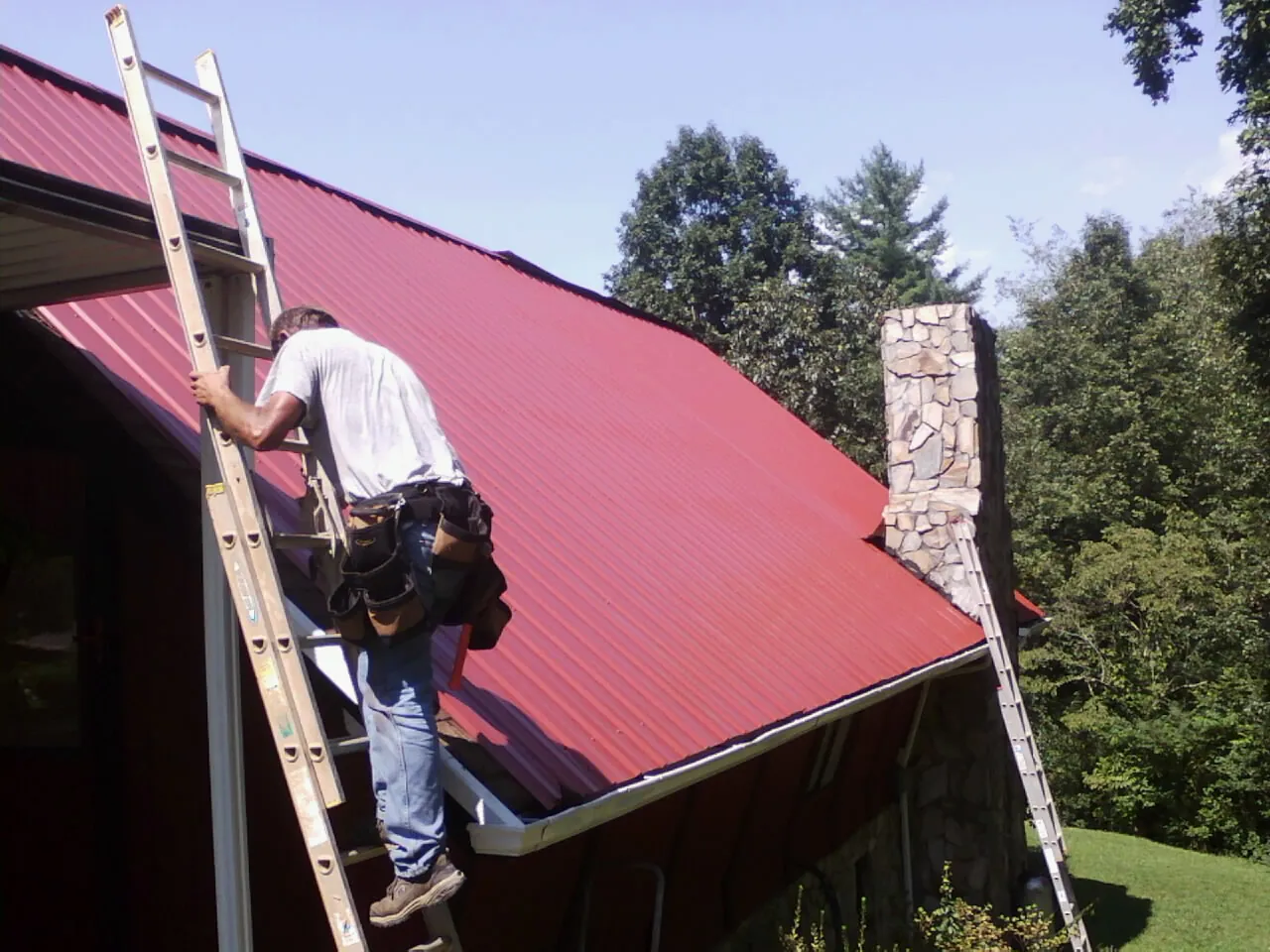 Roofer on a ladder beside a red metal roof and stone chimney — Soffit & Fascia Repair by Roofing Brothers in Lake Geneva